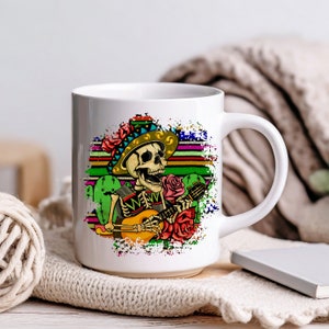 May include: White ceramic mug with a colorful illustration of a skeleton wearing a sombrero and playing a guitar. The background features a colorful striped pattern and cactus plants.
