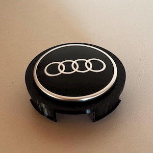 Puede incluir: Tapa central de llanta negra y plateada con el logotipo de Audi. El logotipo presenta cuatro anillos entrelazados blancos sobre un fondo negro. La tapa central de la llanta es redonda con una base de plástico negro.