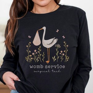 May include: Black long-sleeve shirt featuring a stork carrying a bundle, surrounded by floral and butterfly graphics. The text "womb service surgical tech" is printed below the graphic. The shirt is made of a soft material.