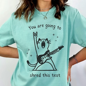 May include: A light teal t-shirt with a black graphic of a cat playing an electric guitar. The text "You are going to shred this test" is above the cat illustration. The shirt has a relaxed fit and short sleeves.