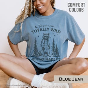 May include: Blue jean colored t-shirt with the text "This year's been TOTALLY WILD BUT WE MADE IT" and a raccoon graphic. The shirt also has the words "COMFORT COLORS" printed on it.