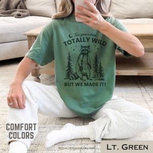 May include: Light green t-shirt with a raccoon graphic and the text "This year's been TOTALLY WILD BUT WE MADE IT!". The shirt is part of the "Comfort Colors" collection.