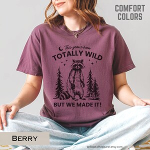 May include: Berry-colored t-shirt with a raccoon graphic and the text "This year's been totally wild, but we made it!" The design includes two pine trees and stars. The shirt is part of the Comfort Colors collection.