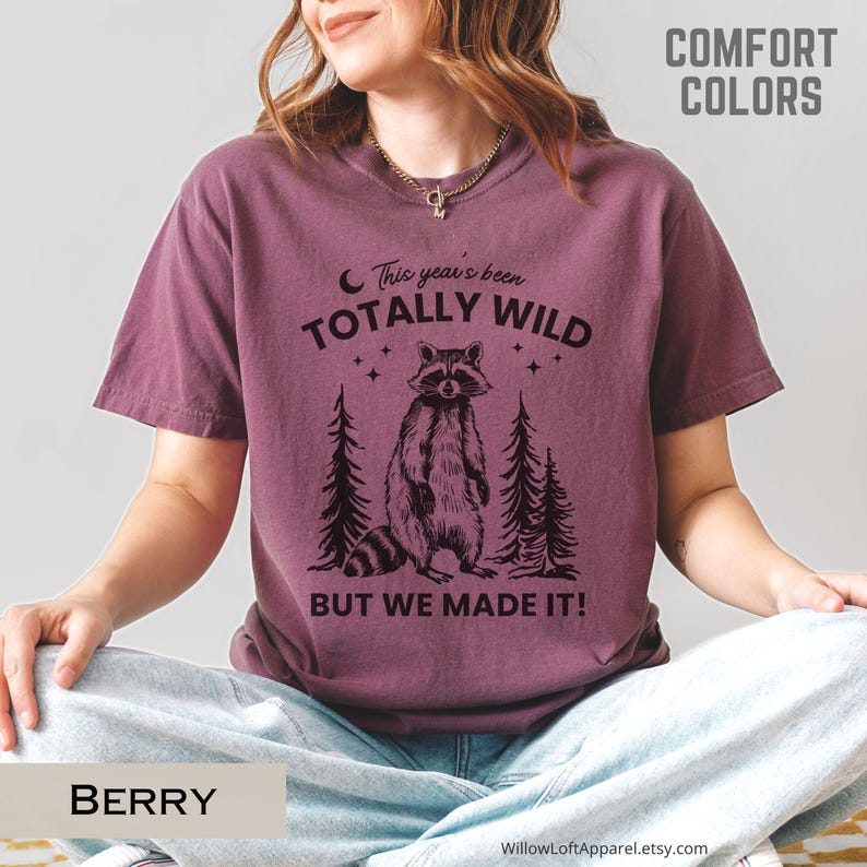May include: Berry-colored t-shirt with a raccoon graphic and the text "This year's been totally wild, but we made it!" The design includes two pine trees and stars. The shirt is part of the Comfort Colors collection.