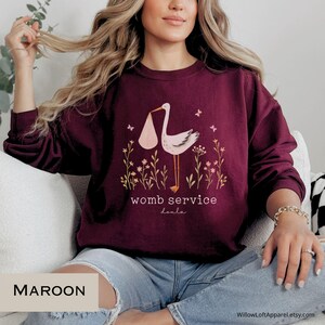 Może przedstawiać: Bordowa bluza z nadrukiem bociana niosącego dziecko w r&oacute;żowym pakunku, z białym napisem "womb service doula". Projekt zawiera kwiatowe i motylkowe akcenty. Okrągły dekolt.