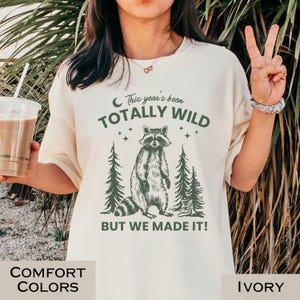 May include: Ivory-colored t-shirt with a graphic of a raccoon standing between two pine trees. The text reads "This year's been TOTALLY WILD BUT WE MADE IT!" in green. The shirt is part of the Comfort Colors collection.