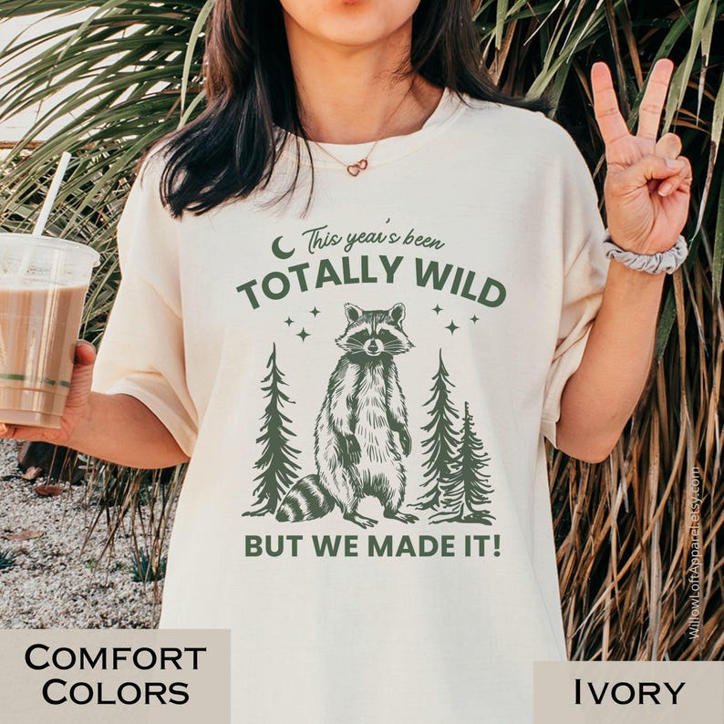 May include: Ivory-colored t-shirt with a graphic of a raccoon standing between two pine trees. The text reads "This year's been TOTALLY WILD BUT WE MADE IT!" in green. The shirt is part of the Comfort Colors collection.
