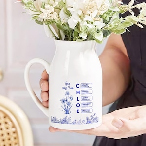 May include: White ceramic pitcher with a handle, filled with white flowers and greenery. The pitcher has blue floral illustrations and text that reads "God says I am" and the name "CHLOE" with associated words.