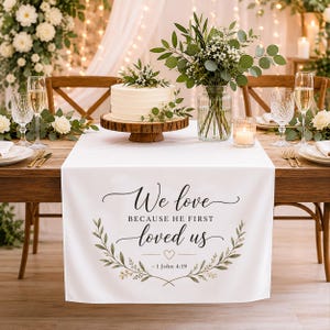 May include: A white rectangular table runner featuring the black printed text We love BECAUSE HE FIRST loved us and 1 John 4:19, framed by two green leafy branches and a small heart icon, centered on a wooden table with a cake and floral centerpiece.