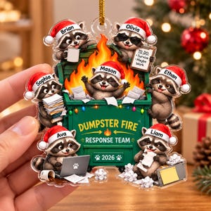 May include: A clear acrylic Christmas ornament featuring six raccoons wearing Santa hats around a green dumpster filled with orange flames. The text reads DUMPSTER FIRE RESPONSE TEAM and 2026. Each raccoon is labeled with names Brian, Olivia, Mason, Sophia, Ethan, Ava, and Liam.