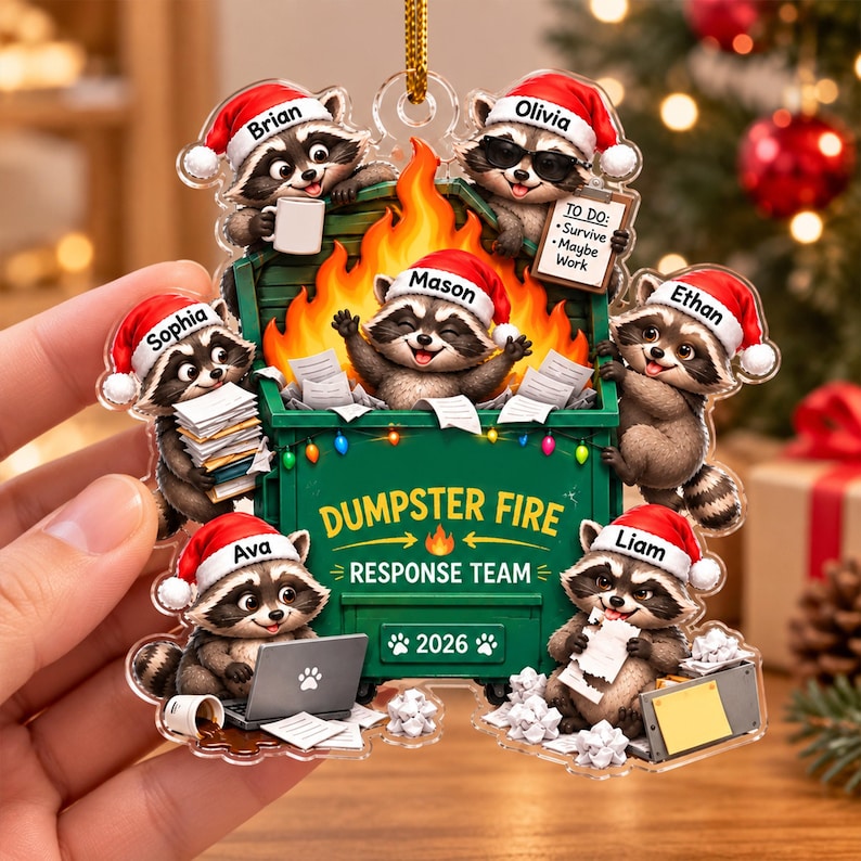 May include: A clear acrylic Christmas ornament featuring six raccoons wearing Santa hats around a green dumpster filled with orange flames. The text reads DUMPSTER FIRE RESPONSE TEAM and 2026. Each raccoon is labeled with names Brian, Olivia, Mason, Sophia, Ethan, Ava, and Liam.