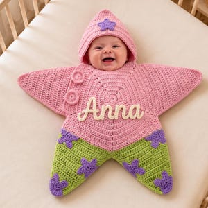 May include: A pink and green star-shaped crochet baby swaddle blanket with a hood. The top is pink with a purple star on the hood and two circular buttons. The bottom is green with purple star appliques. The name Anna is stitched in white.