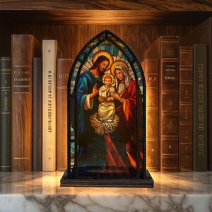 May include: A stained glass panel depicting the Holy Family, with Jesus, Mary, and the infant Jesus. The artwork features vibrant colors and is set in a dark wooden frame. The panel is displayed on a marble surface, surrounded by antique books.