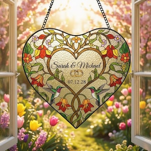 May include: Heart-shaped stained glass window hanging with a floral design, hummingbirds, and the names "Sarah & Michael" with a date. The window is framed by a black chain and set against a blurred garden background.