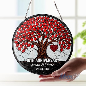 May include: Circular stained glass artwork depicting a tree with red leaves and a heart. The text reads "40th Anniversary" with names and a date. The piece is framed in black and suspended by a chain.