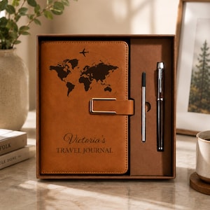 May include: A tan leather-like travel journal with a world map graphic, a small airplane icon, and the text "Victoria's TRAVEL JOURNAL" embossed on the cover. The journal features a gold-toned metal clasp and is presented in a box with a matching pen.