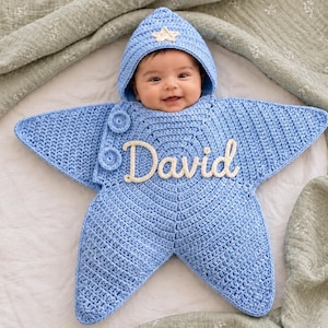 May include: A light blue crocheted star-shaped baby swaddle blanket with a hood featuring a small cream-colored star appliqué. The front displays the name David in cream-colored cursive lettering and includes two circular crocheted buttons.