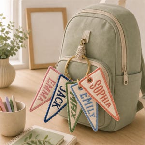May include: A sage green backpack featuring a gold metal ring attached to the front zipper. Five triangular fabric pennants hang from the ring, each embroidered with a name: EMMA, JACK, MAX, EMILY, and SOPHIA, in various thread colors.
