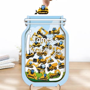 Op de afbeelding: Een lichtblauwe houten beloningspot in de vorm van een weckpot met de tekst James's Reward-Jar. De binnenzijde is gevuld met illustraties van gele bouwvoertuigen, zoals graafmachines en kiepwagens, inclusief een bijpassend geel bulldozer-onderdeel.