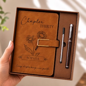 May include: A tan notebook with a gold metal clasp, featuring the text Chapter THIRTY, the name SOPHIA, and the phrase Every chapter brings new growth, alongside a floral line drawing. The set includes a matching pen and a refill in a box.