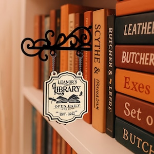 Puede incluir: Un letrero de biblioteca de metal negro con el texto "LEANOR'S LIBRARY" y "OPEN DAILY EST. 2022" cuelga de un soporte decorativo. El letrero está suspendido por cadenas frente a una estantería llena de libros.
