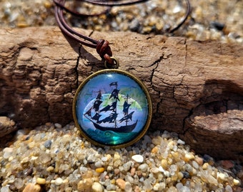 Op de afbeelding: Een ketting van bruin lederen koord met een ronde glazen hanger met een zwart silhouet van een piratenschip met zeilen tegen een blauwe en witte achtergrond.