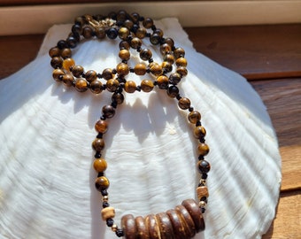 May include: A brown and gold tiger's eye beaded necklace with a wooden pendant. The necklace is displayed on a white seashell.