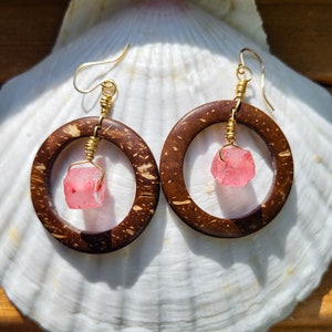 Peut inclure: Une paire de boucles d'oreilles avec des perles de verre roses et des cercles en coquille de noix de coco brune. Les boucles d'oreilles sont suspendues à un fil d'or.
