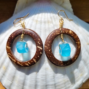 Op de afbeelding: Een paar oorbellen met een bruine houten cirkel en een blauwe glazen traanvormige hanger. De oorbellen hangen aan een gouden draad.