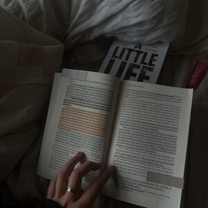 May include: A close-up of an open book with the title "A Little Life" visible on the cover. The book is resting on a bed with a hand holding a ring on the page.