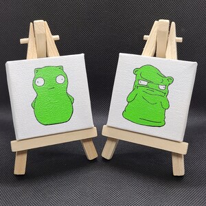 May include: Two small green cartoon characters painted on canvases, each mounted on a wooden easel. The character on the left is standing upright with a surprised expression. The character on the right is hunched over with a grumpy expression.