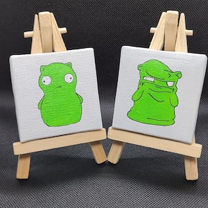 May include: Two small green cartoon characters painted on canvases, each mounted on a wooden easel. The character on the left is a green blob with eyes and a mouth. The character on the right is a green blob with a face and arms.