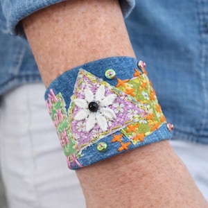 Puede incluir: Pulsera de puño de mezclilla con patrones florales y geométricos bordados. La pulsera presenta una flor blanca con un centro negro, rodeada de acentos rosas, verdes y naranjas. Adornada con pequeñas cuentas de colores.