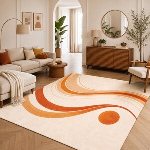 May include: Cream-colored area rug with an abstract design in shades of orange and peach. The rug features a wavy pattern and a large orange circle. The rug is placed in a living room with a sofa, coffee table, and wooden furniture.