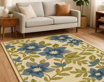 Puede incluir: Una alfombra rectangular con diseño floral. Presenta grandes flores azules con centros blancos y hojas verdes sobre un fondo crema. El borde es de un verde más oscuro.
