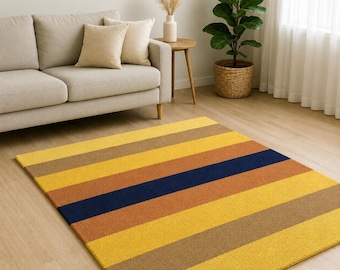 May include: A rectangular area rug featuring horizontal stripes in shades of mustard yellow, burnt orange, dark navy blue, and light brown. The rug has a textured, low-pile surface and is positioned on a light-colored wooden floor in a room.