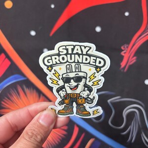 May include: A sticker featuring a cartoon electrical plug character wearing sunglasses and holding tools. The sticker has the text "STAY GROUNDED" in a white bubble. The character is wearing a tool belt and brown boots.