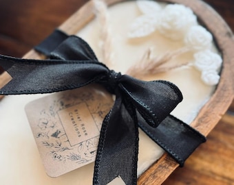 May include: A decorative candle in a wooden bowl, featuring a creamy white wax with floral accents and a black bow. The candle has a tag that reads "Brier Rose Creations". The bowl is oval-shaped and made of wood.