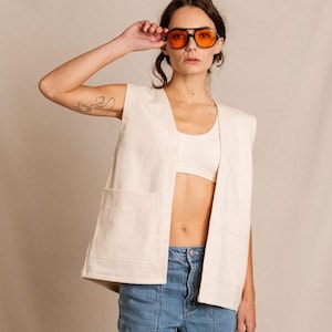 May include: A woman wearing a white sleeveless utility vest over a white tank top, blue jeans, and white sneakers. She is wearing orange-tinted sunglasses.