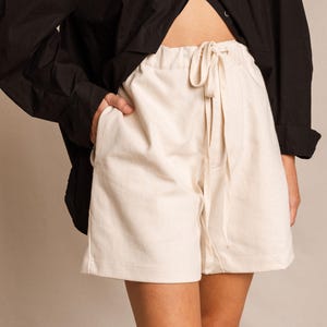 Puede incluir: Pantalones cortos color crema con cintura con cordón y bolsillos. Los pantalones cortos están hechos de una tela suave y ligera. Una camisa negra abotonada es parcialmente visible en el fondo.
