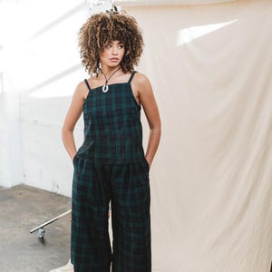 May include: A woman wearing a green and black plaid matching set, consisting of a square-neck tank top and wide-leg pants. The outfit is paired with black boots and a silver necklace. The background is a neutral color.