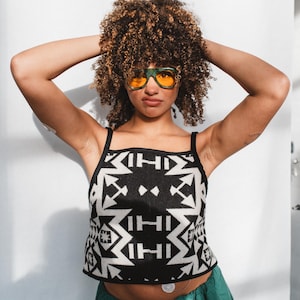 May include: A black and white patterned crop top with a geometric design. The top has thin straps and is paired with green pants. The person is wearing orange-tinted sunglasses.