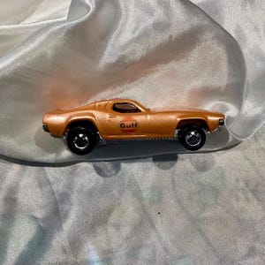 May include: A die-cast, orange toy car with black wheels. The car has the word "Gulf" printed on the side in a circular logo. The car is a vintage model, likely from the 1970s or 1980s.