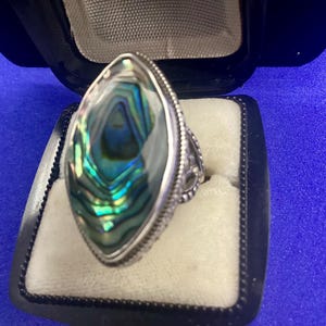May include: Abalone shell ring with a silver band. The ring features an oval-shaped abalone shell with iridescent green, blue, and gold hues. The silver band has a detailed, ornate design. The ring is displayed in a black jewelry box with a white interior.