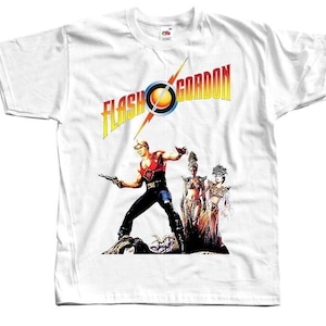 Può includere: Maglietta bianca con una stampa grafica del film "Flash Gordon". La grafica presenta un uomo in un costume rosso e nero che tiene una pistola, e due donne in costumi colorati.