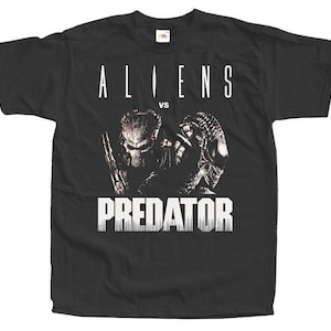 Può includere: T-shirt nero con la scritta bianca "ALIENS vs PREDATOR". La grafica raffigura i personaggi Alien e Predator in bianco e nero. La maglietta è a girocollo.