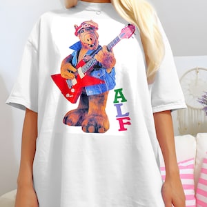 May include: White oversized t-shirt with a graphic print of the character ALF playing a red electric guitar. The text "ALF" is printed vertically in rainbow colors below the image.