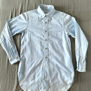 Puede incluir: Una camisa de manga larga de color azul claro con un botón y un bolsillo en el pecho. La camisa tiene un dobladillo curvo y un cuello abotonado.