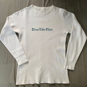 以下が含まれることがあります： リブのついた白い長袖サーマルシャツ。クルーネックで、前面にダークグリーンで「Times Like These」という文字が刺繍されています。