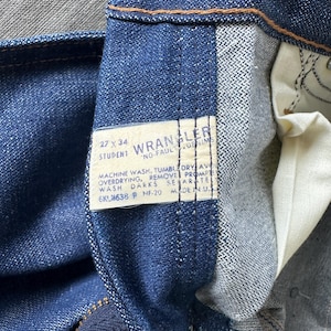 Op de afbeelding: Close-up van een blauwe spijkerbroek met een zichtbaar label. Het label luidt "WRANGLER" en bevat wasvoorschriften en maat 27 x 34. De jeans is gemaakt in de VS.